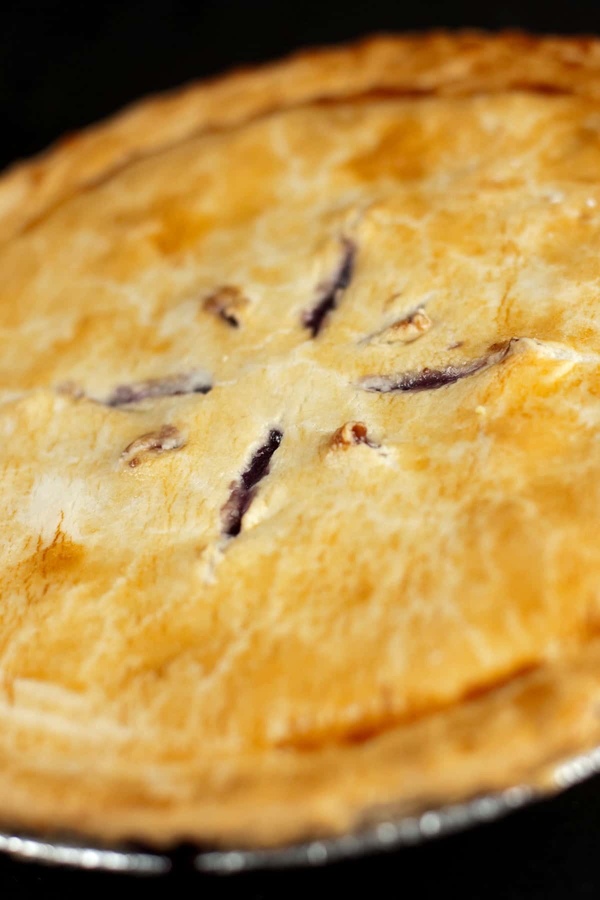 A whole 2-crust pie, against a black background. Slits cut into the top crust reveal it to be a blueberry pie.