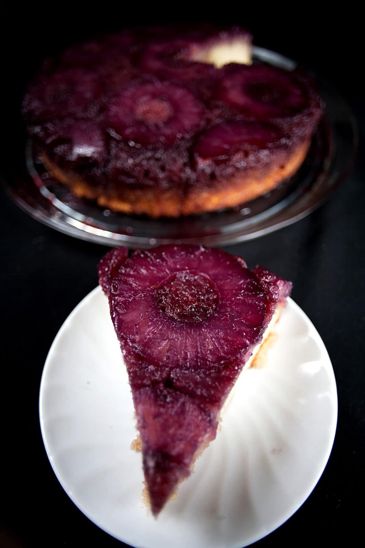 French Martini Upside Down Cake - A round pineapple upside down cake, with a VERY purple top! A slice has been cut out and is on a plate next to the main cake.