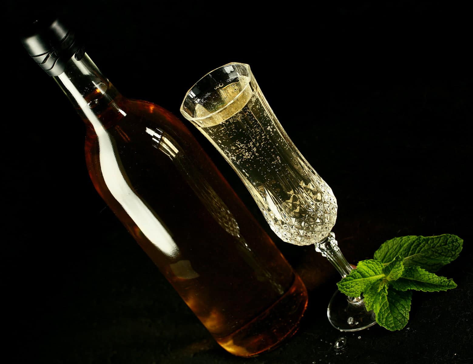 A full wine bottle and a glass of mint wine. There is a spring of mint at the base of the glass, everything is against a black background.