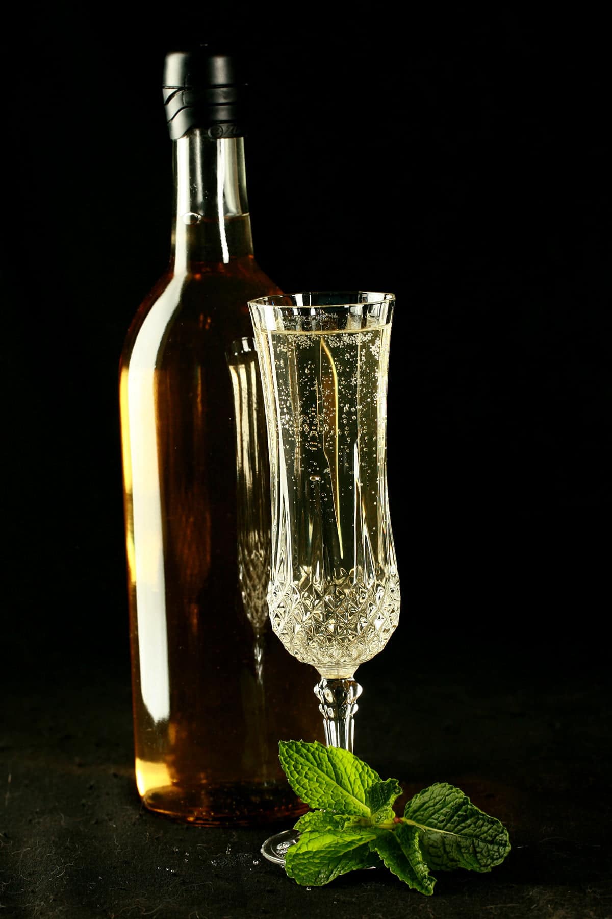 A full wine bottle and a glass of mint wine. There is a spring of mint at the base of the glass, everything is against a black background.