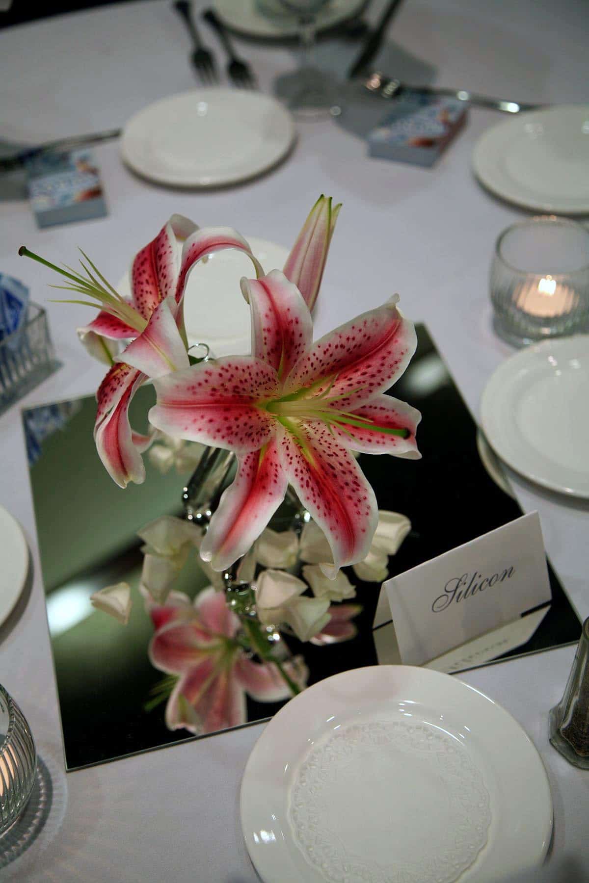 2 stargazer lilies and a bud, in a tripod vase made from test tubes.  The arrangement rests on a large square mirror tile.