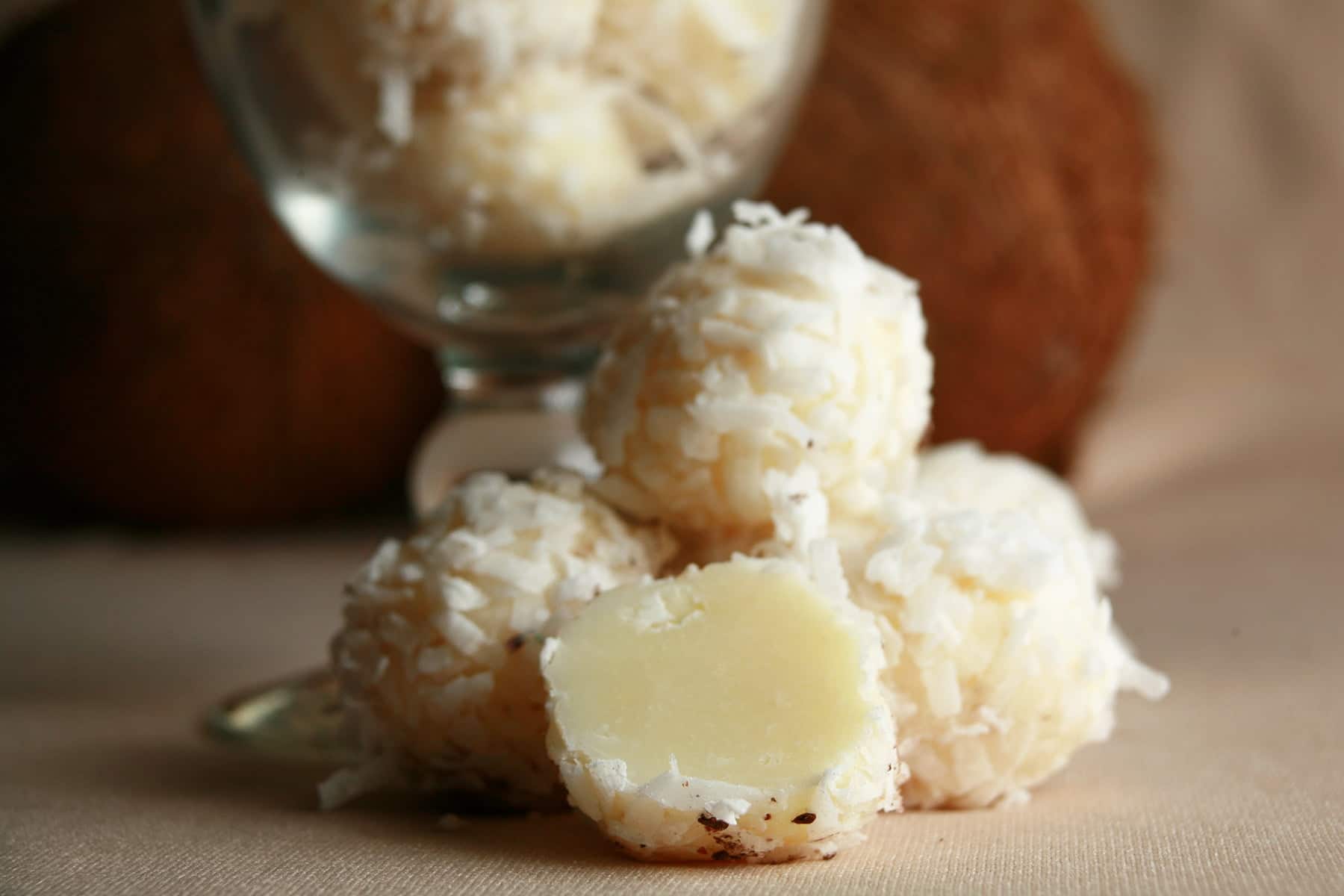 Several Tropical White Chocolate Truffles are piled at the base of a cocktail glass. More truffles are contaied within the glass, and there is a coconut behind it.