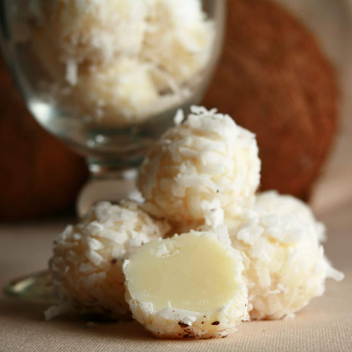 Several Tropical White Chocolate Truffles are piled at the base of a cocktail glass. More truffles are contained within the glass, and there is a coconut behind it.