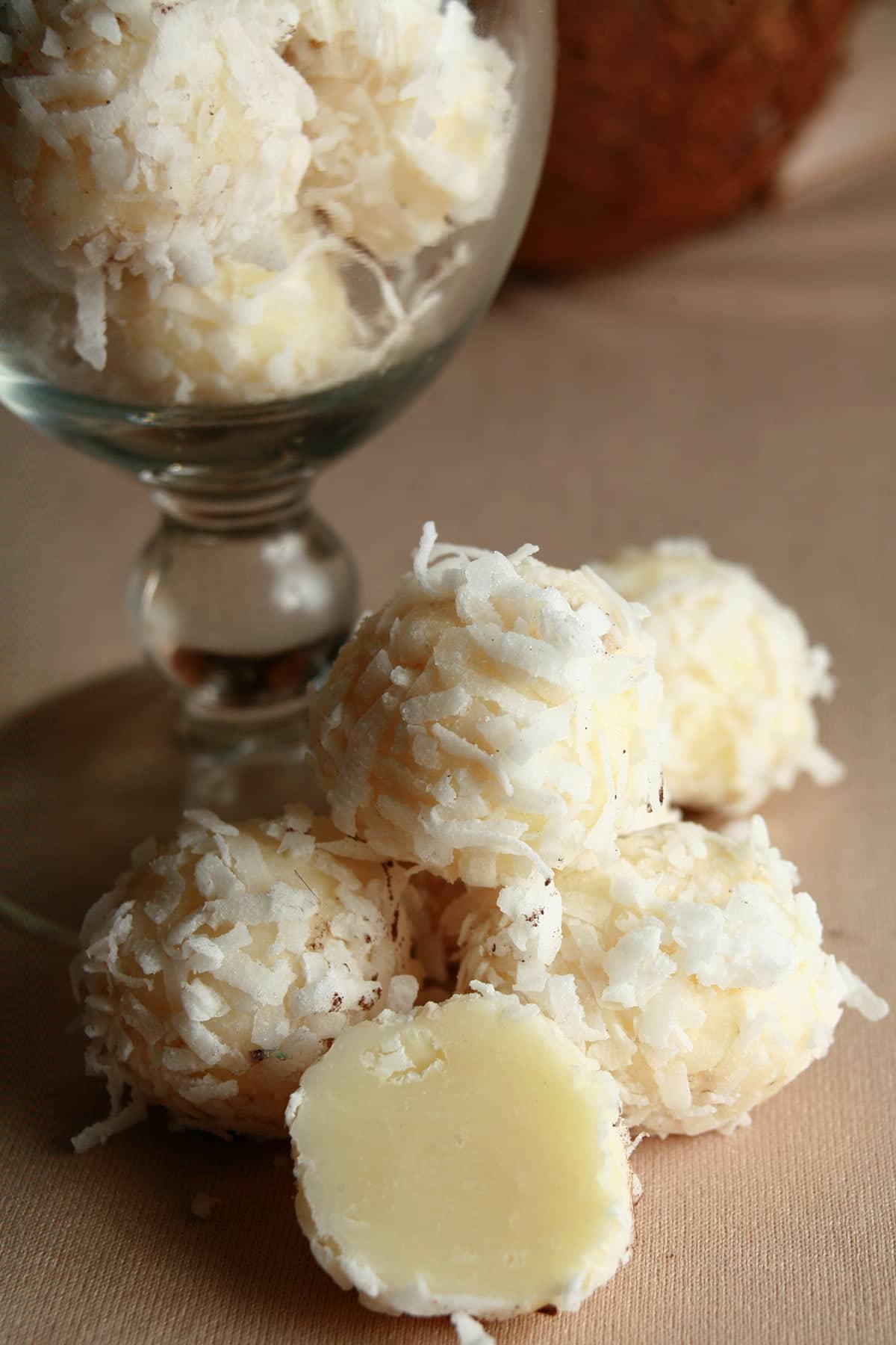 Several Coconut Covered White Chocolate Truffles are piled at the base of a cocktail glass. More truffles are contained within the glass, and there is a coconut behind it.