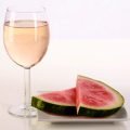 A glass of pale pink wine, next to a white plate with watermelon slices on it.