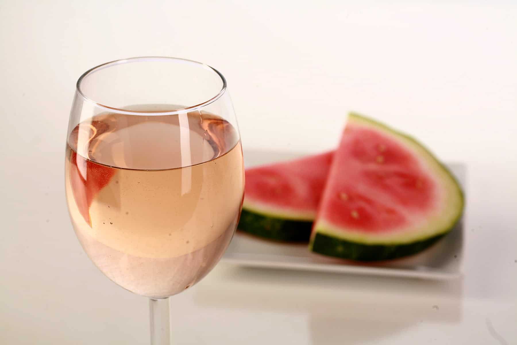 A glass of pale pink wine, next to a white plate with watermelon slices on it.