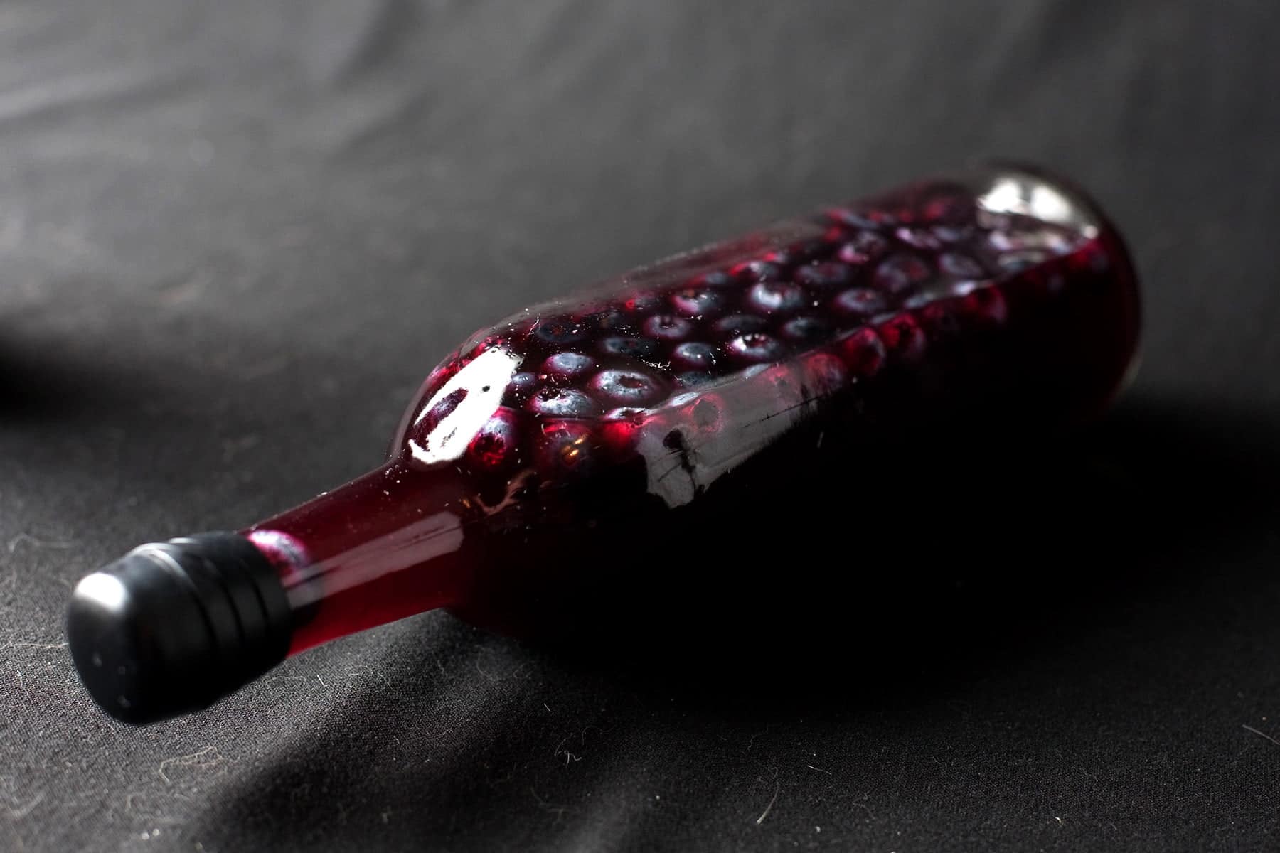 A wine bottled filled with dark purple liqueur and floating blueberries is pictured.