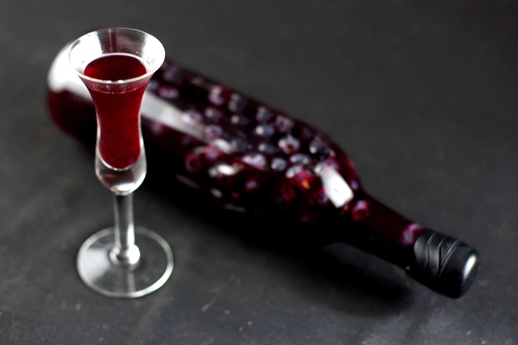 A wine bottled filled with dark purple liqueur and floating blueberries is pictured next to a fluted shot glass filled with the dark purple liquid.