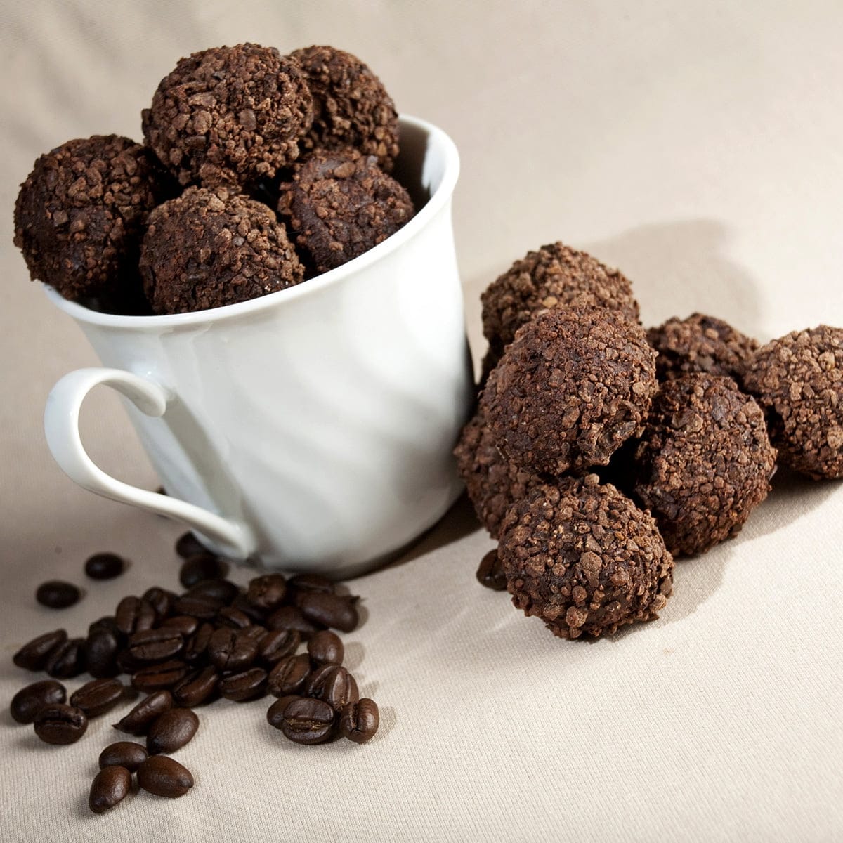 Dark chocolate truffles - coated in smashed coffee beans - fill a white coffee mug. There are stray dark chocolate coffee truffles and coffee beans at the base of the mug.