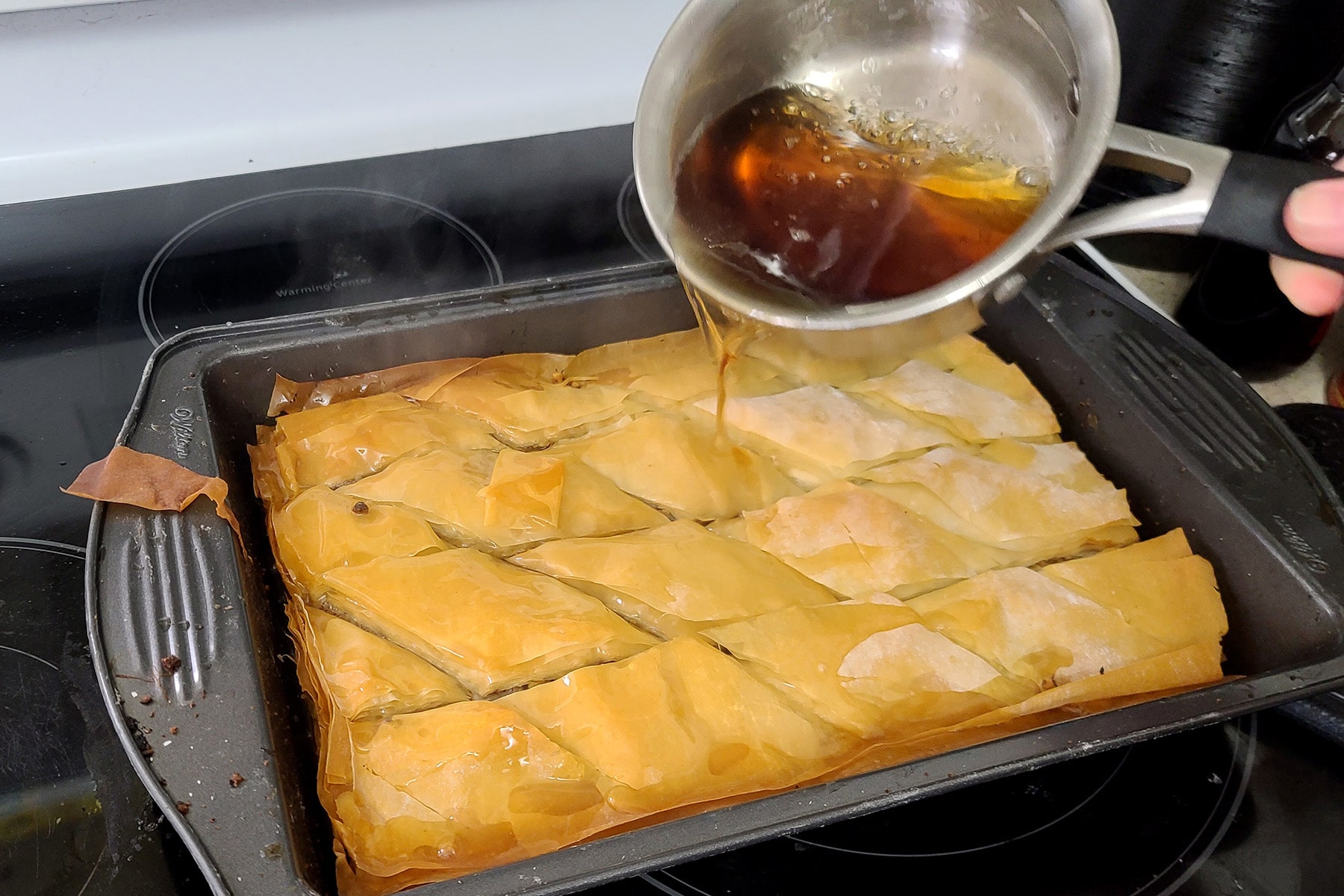 The pot of maple syrup mixture is being poured over the baked pan of baklava.