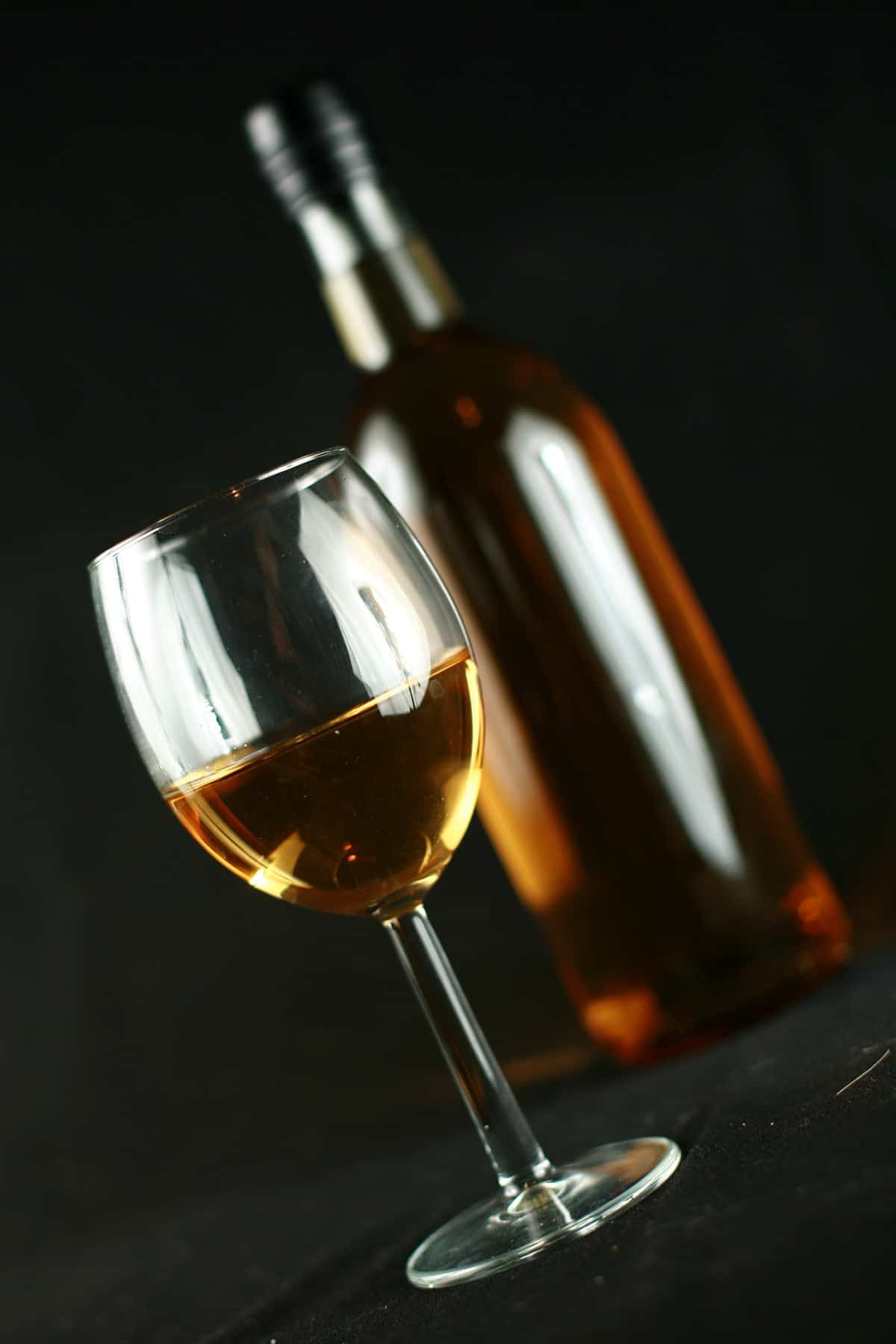 A wine glass and a wine bottle are filled with golden banana wine, against a black background.