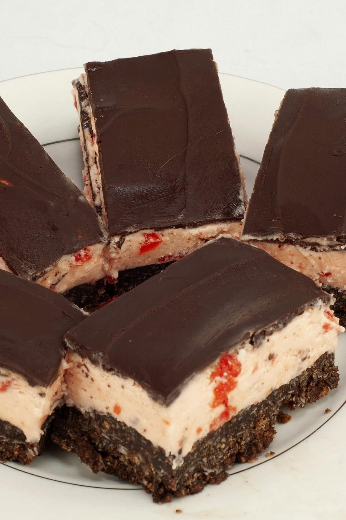 Close up view of a plate of 3 layered bars. The bottom layer looks like a brownie, the middle is a thick pink buttercream with bits of cherry visible, and the top is a smooth chocolate ganache. Maraschino Cherry Nanaimo Bars!