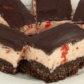 Close up view of a plate of 3 layered bars. The bottom layer looks like a brownie, the middle is a thick pink buttercream with bits of cherry visible, and the top is a smooth chocolate ganache.