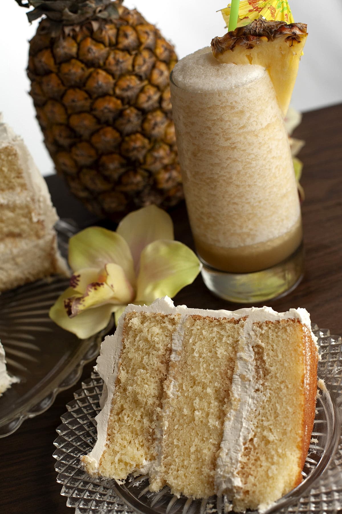 A slice of Bahama Mama torte is shown next to the cake it is cut from. 3 layers of a white cake, filled and frosted with a white frosting. A pimeapple, a yellow cymbidium orchid, and a tropical cocktail - a Bahama Mama - are shown next to and behind the cake.