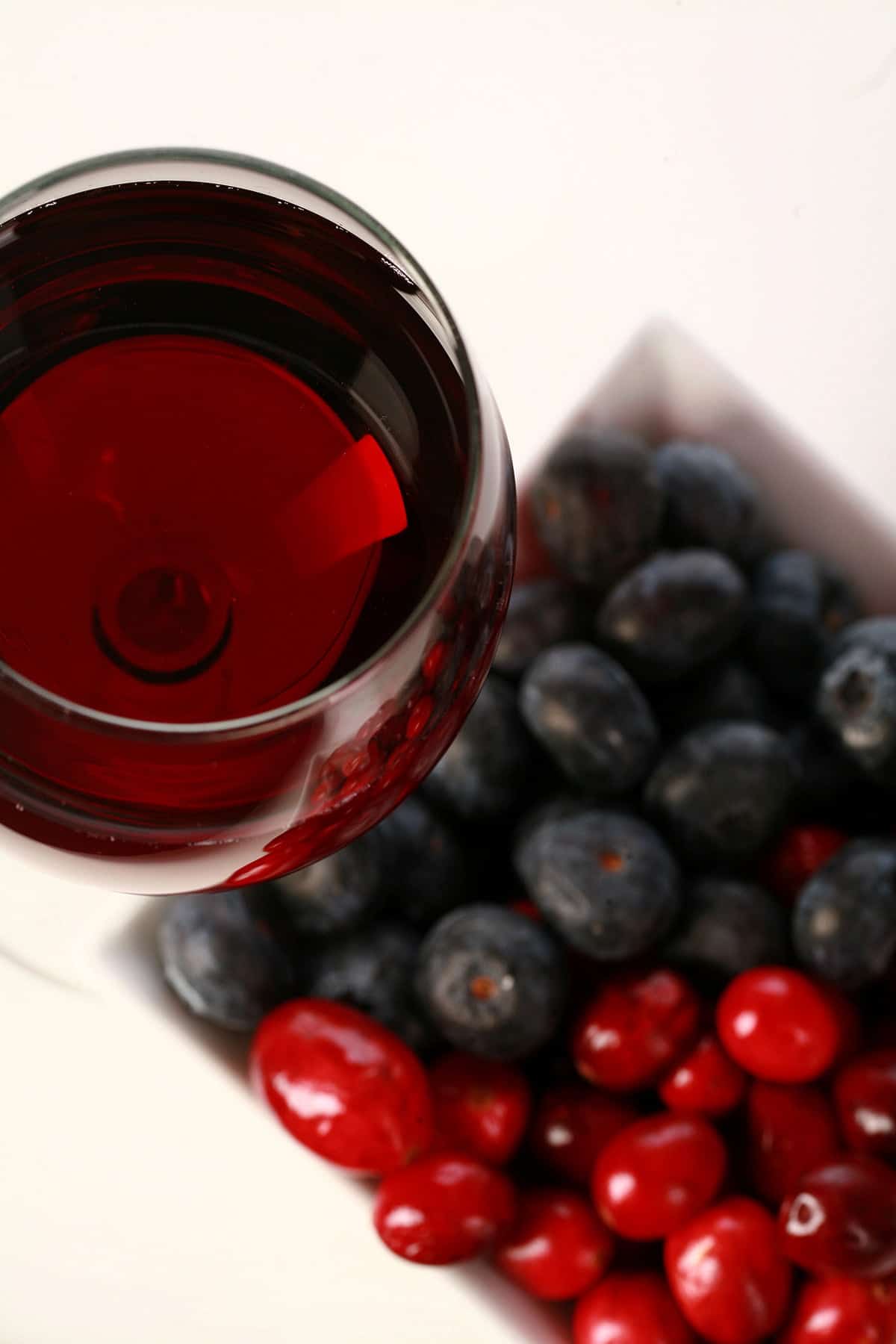 A glass of red wine - made from this faux lingonberry wine recipe - is pictured next to a small bowl of cranberries and blueberries