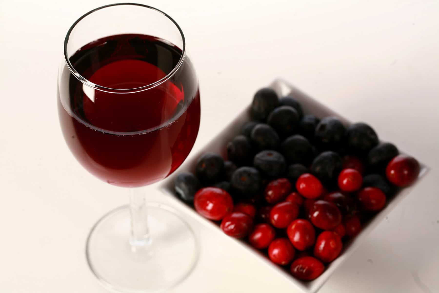 A glass of red wine is pictured next to a small bowl of cranberries and blueberries.