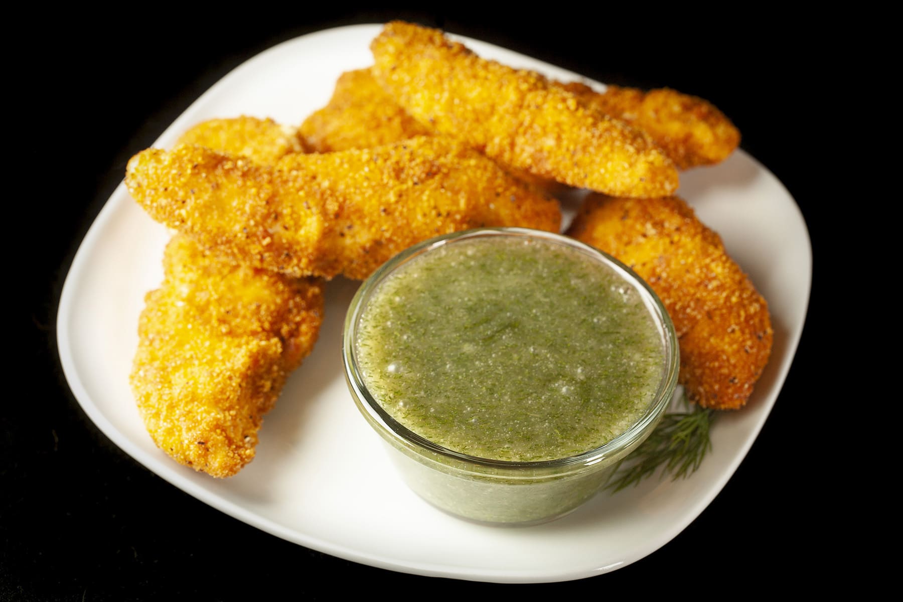 A square shaped white plate with several golden brown chicken tenders on it, along with a glass bowl full of honey dill sauce.