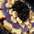 A close up view of a rum runner trifle, in a large glass bowl. Layers of cake cubes, purple pudding, bananas, and blackberries are visible.