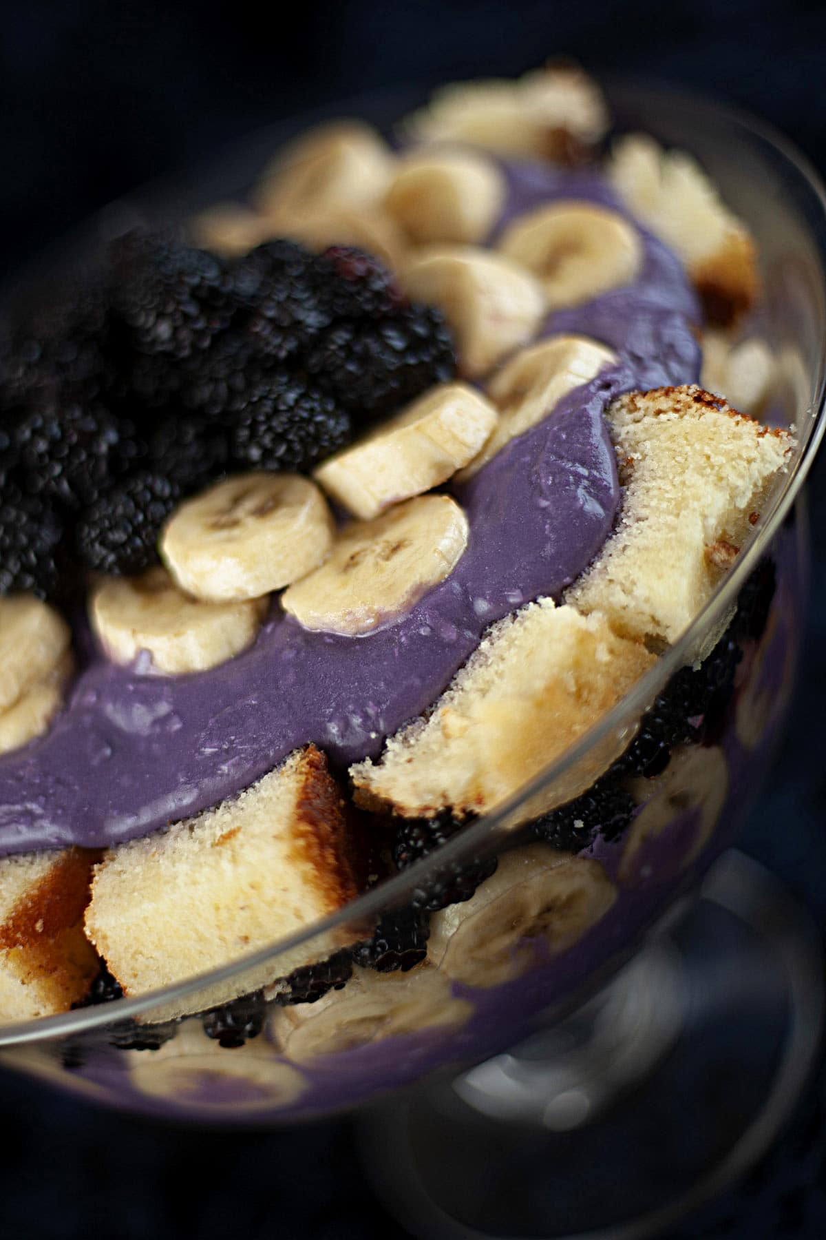 A close up view of a rum runner trifle, in a large glass bowl. Layers of cake cubes, purple pudding, bananas, and blackberries are visible.