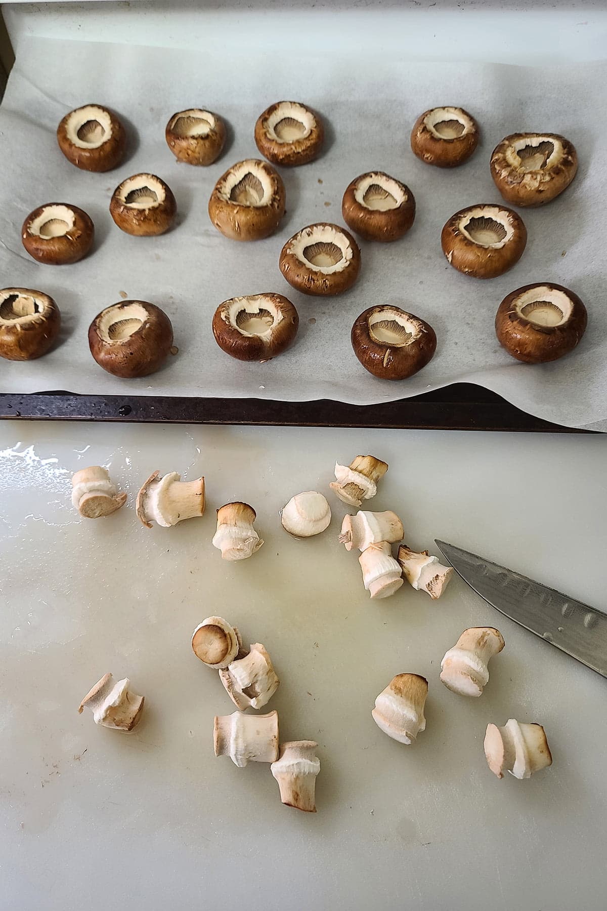 Mushroom caps arramged on a baking sheet, behind a cutting board with mushroom stems on it.