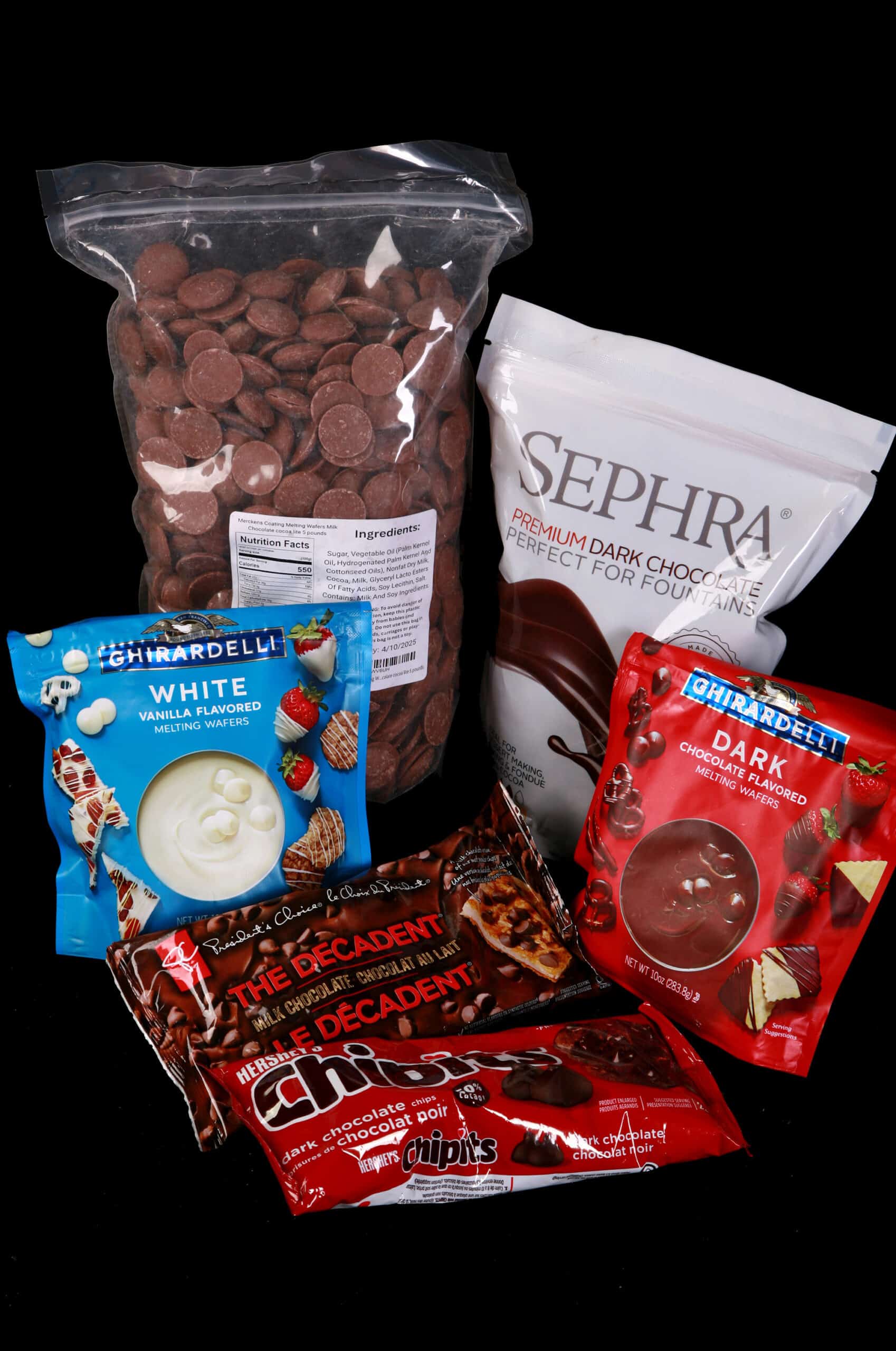 Several bags of chocolate for chocolate fountains, including a big bag of merckens chocolate wafers, Sephra Premium Dark Chocolate, Ghirardelli Wafers, and chocolate chips.