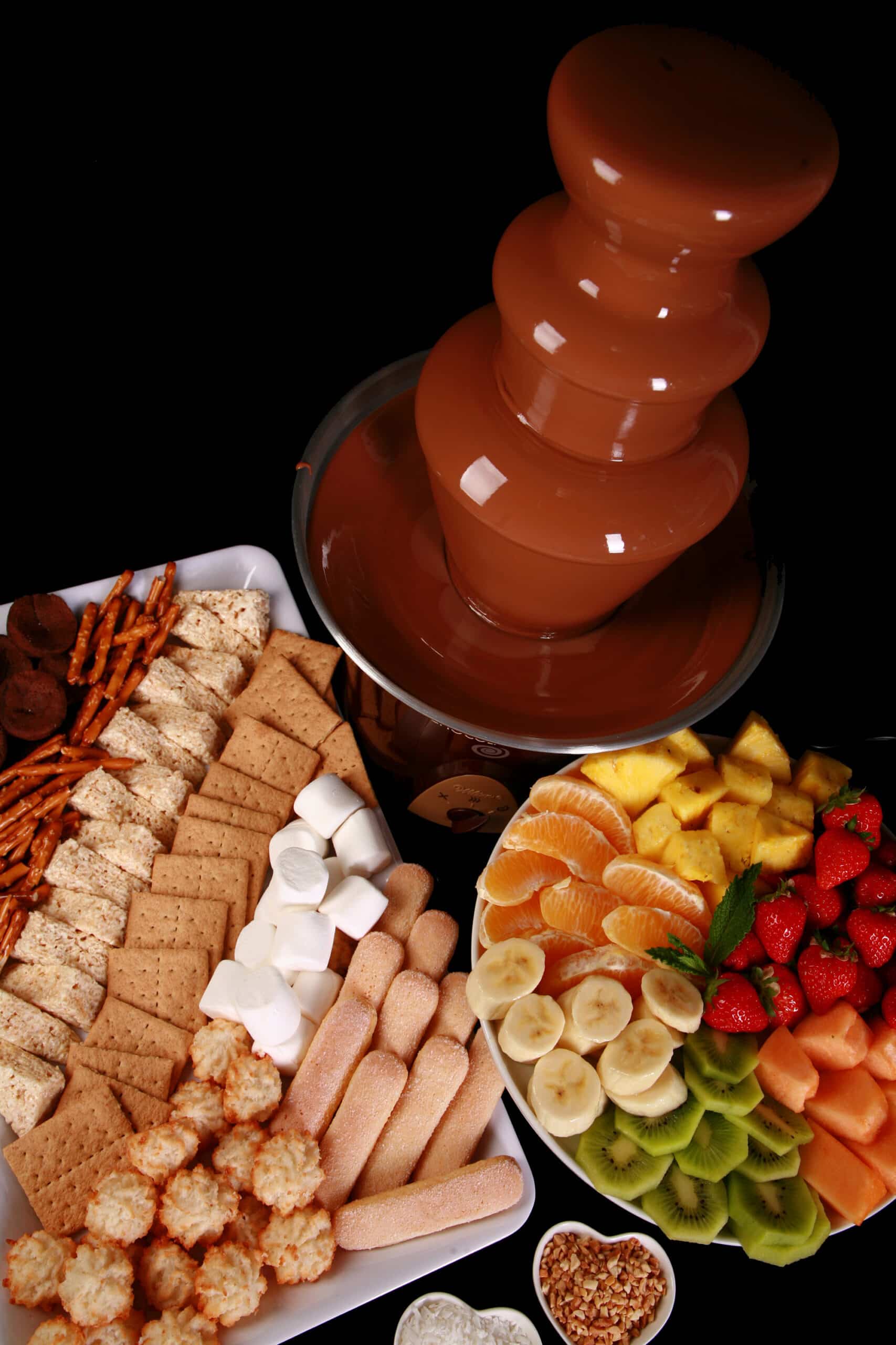 An operating chocolate fountain, with a tray of cookies, marshmallows, pretzels, coconut macaroons and more. Next to it is a raised cake plate with prepared fruit, including bananas, strawberries, pineapple, oranges, and kiwifruit.