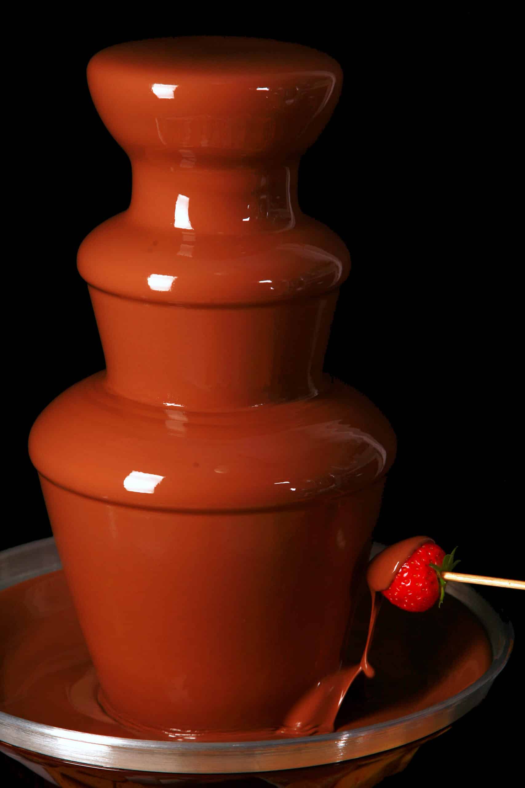 A strawberry on a skewer, being dipped into a chocolate fountain.