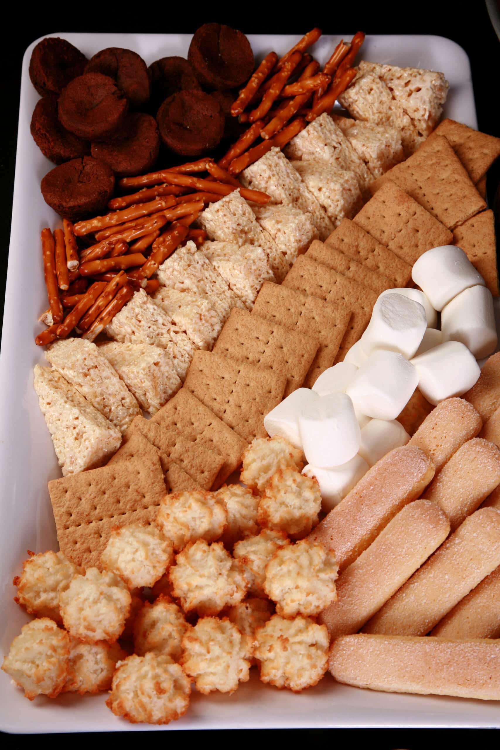 A large display tray of items to dip in a chocolate fountain, including 2 bite brownies, pretzel rods, rice krispie squares, graham crackers, marshmallows, coconut macaroons, and ladyfingers.