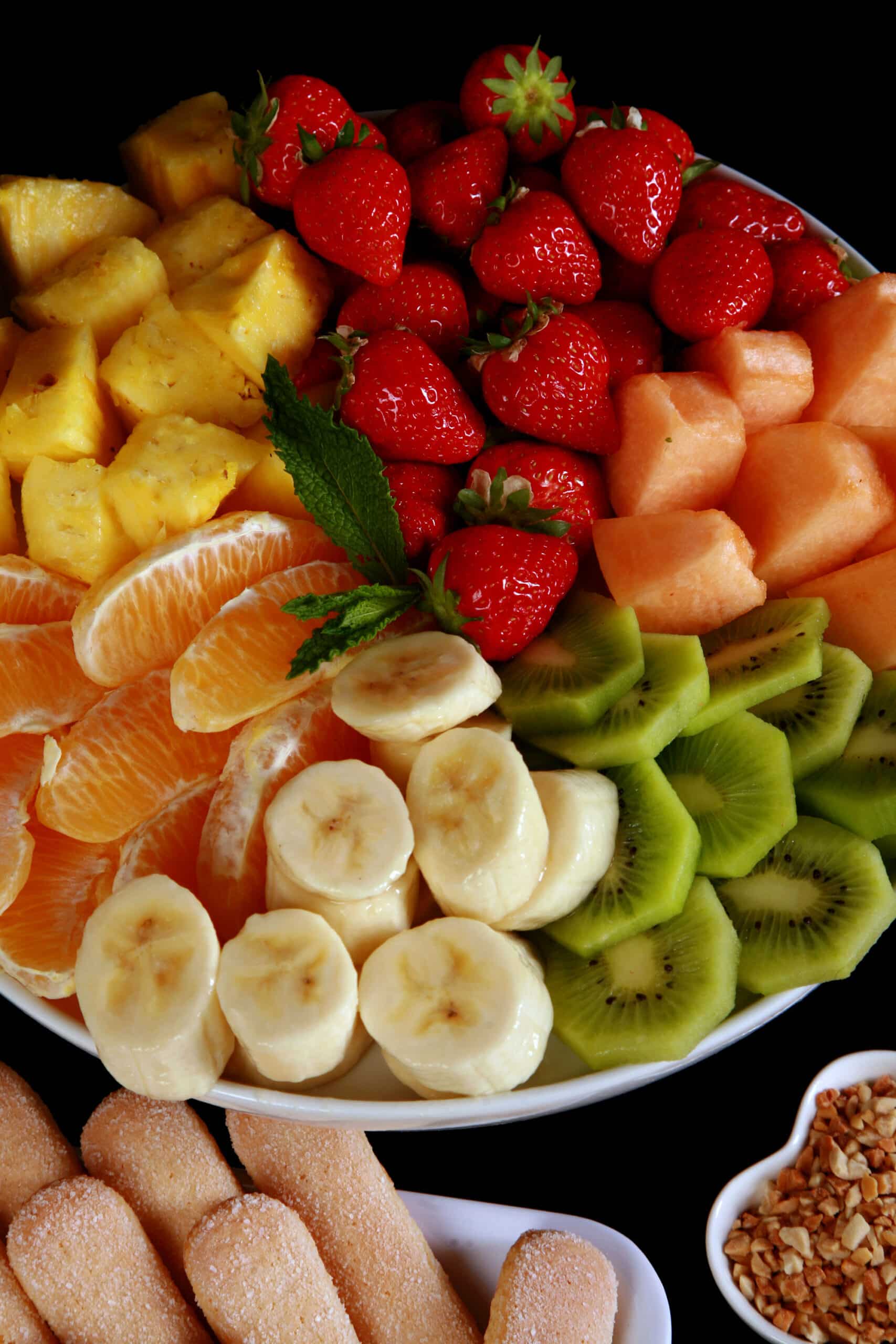 A close up view of a large platter of fruit for dipping in a chocolate fountain. There are strawberries, orange slices, kiwi slices,  banana slices, and chunks of pineapple and cantaloupe.