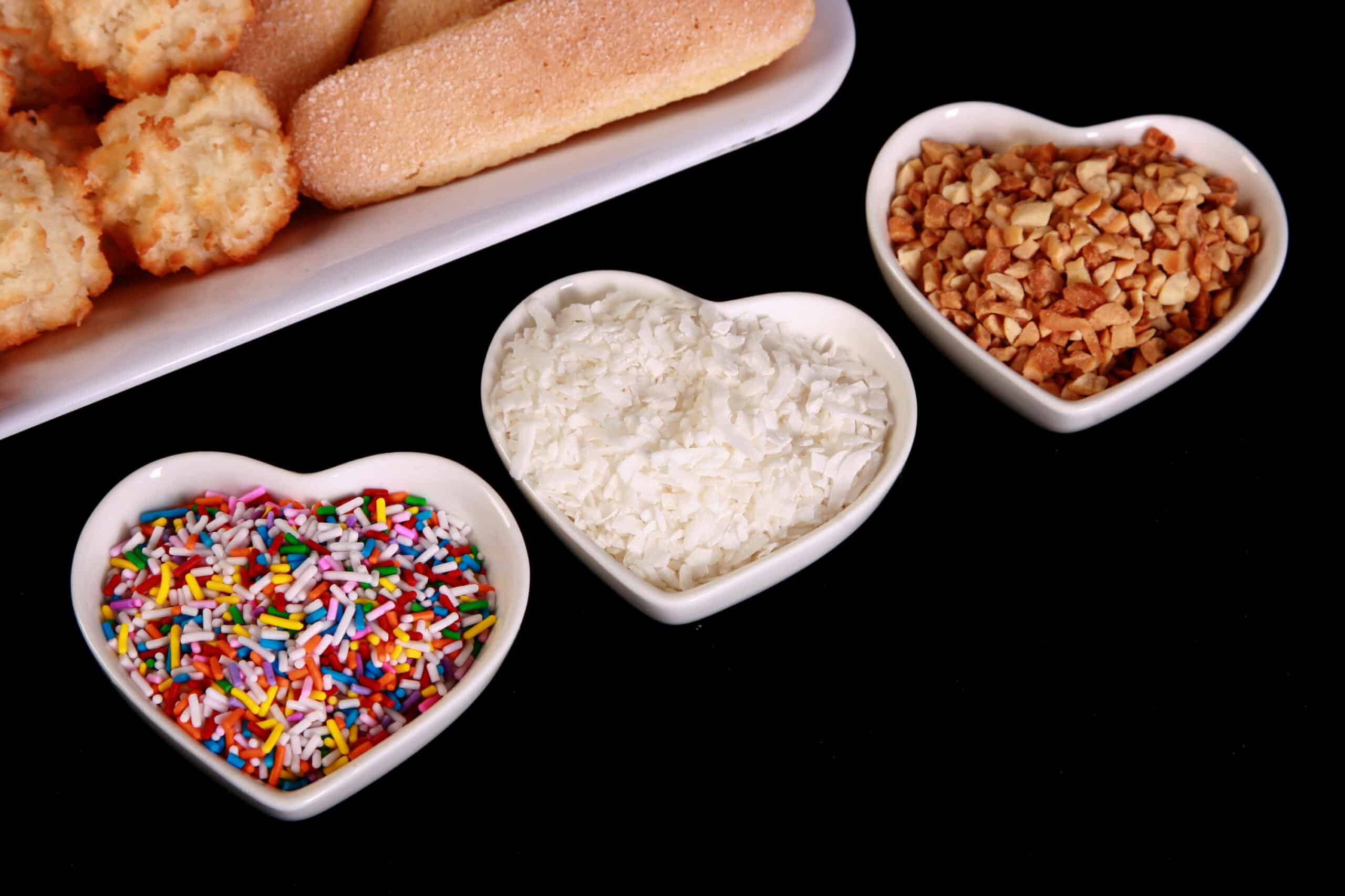 A close up view of 3 little heart shaped ramekins. One has sprinkles, another has coconut flakes, and the third has chopped peanuts.