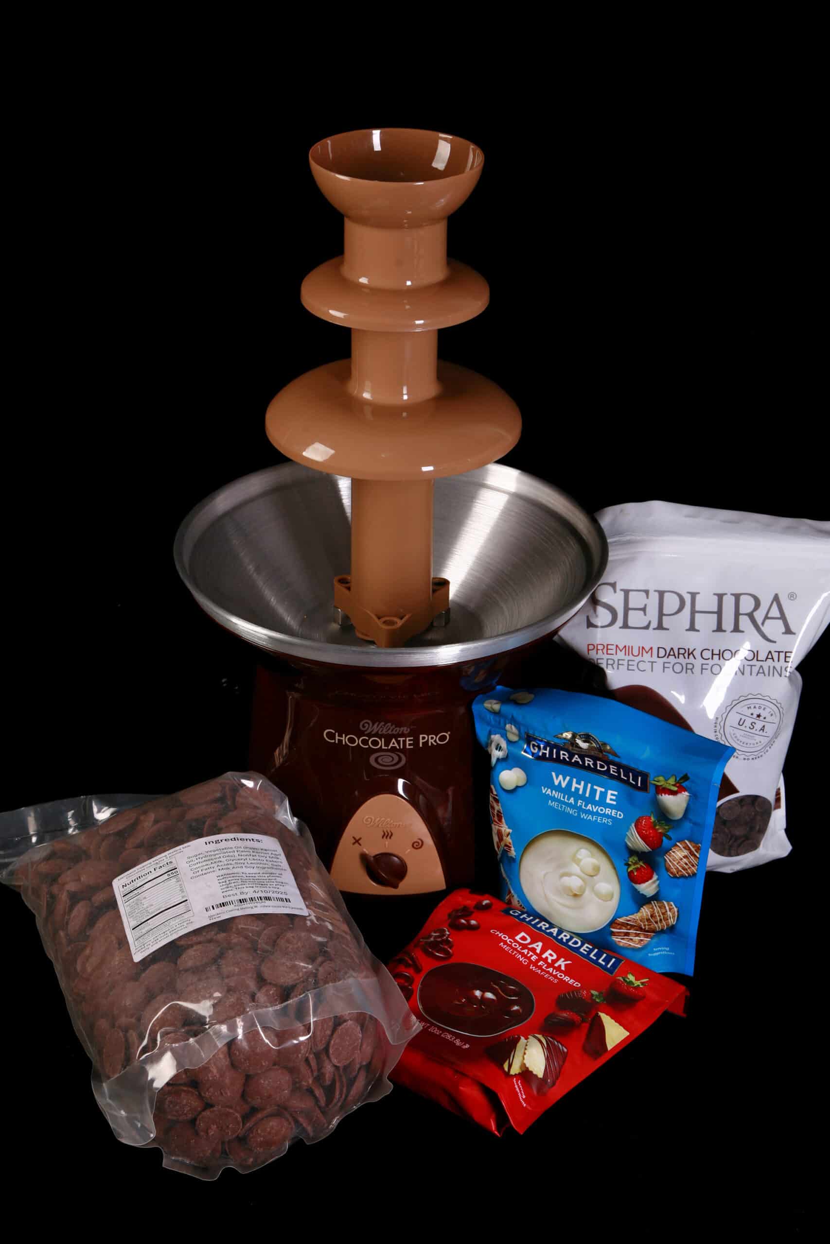 An empty chocolate fountain - the Wilton Chocolate Pro - with various bags of chocolate at the base.  There is Sephra chocolate, Merckens Chocolate Wafers, and two bags of Ghirardelli.