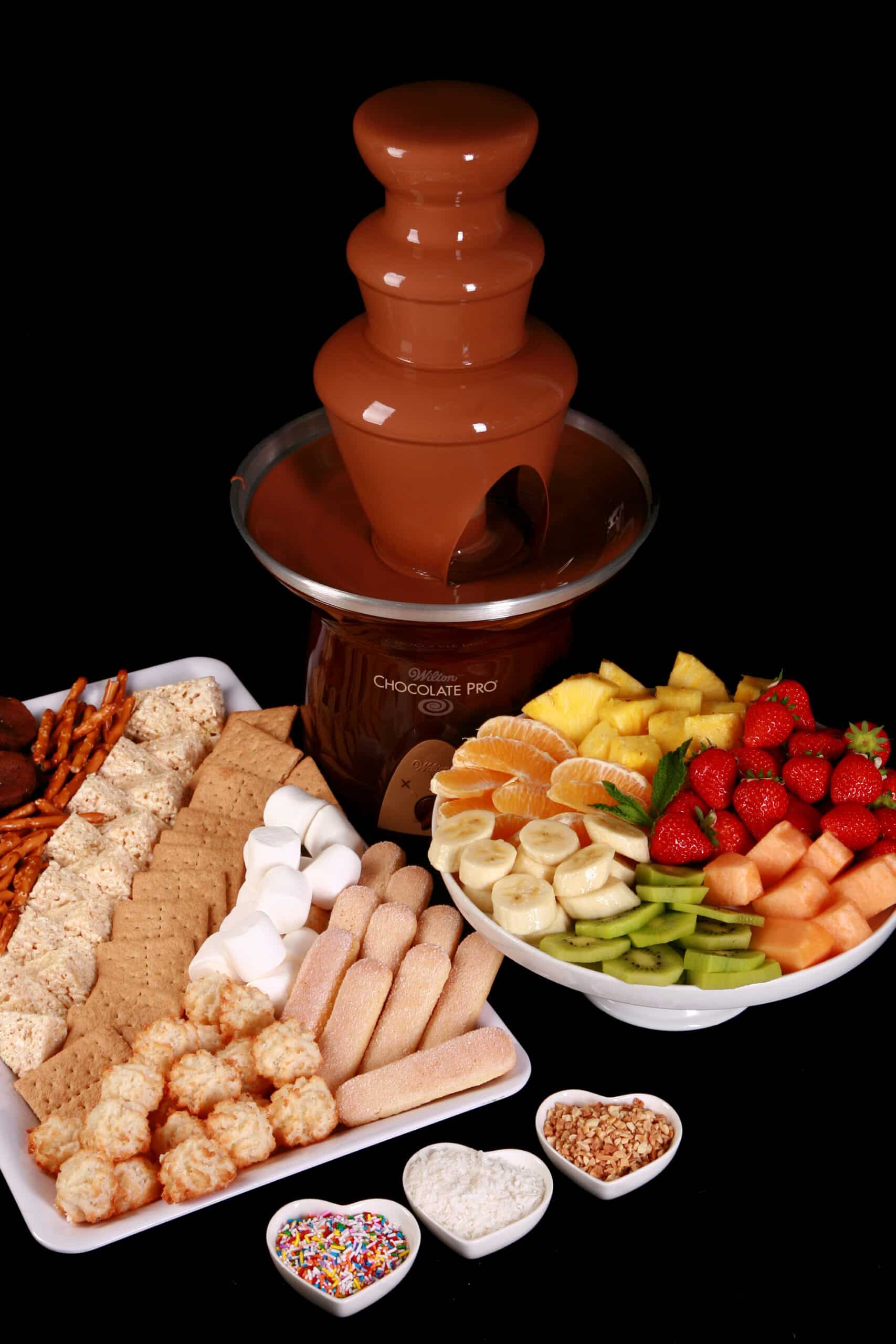 A chocolate fountain with trays of cookies and fruit in front of it.