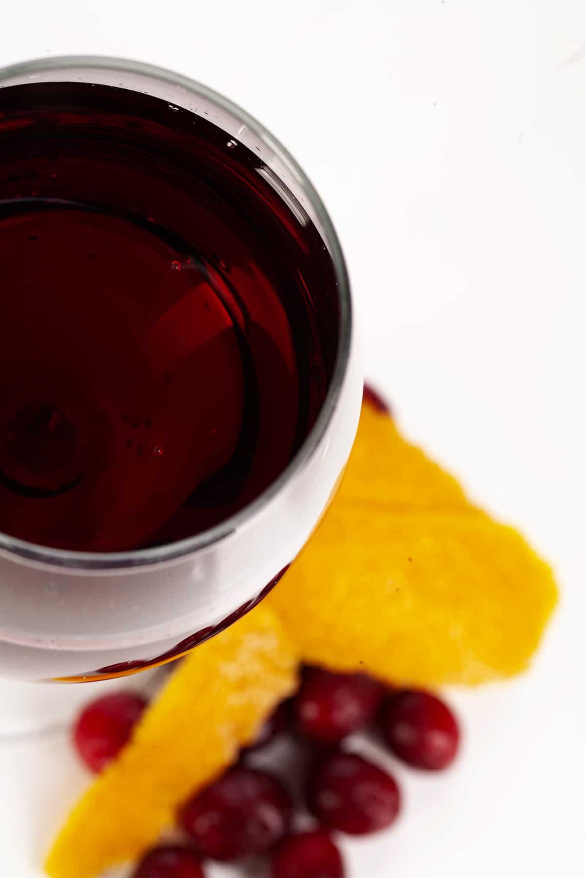 A close up view of a wine glass with with a deep red wine. There are cranberries and orange peels at the base of the glass, against a white background.