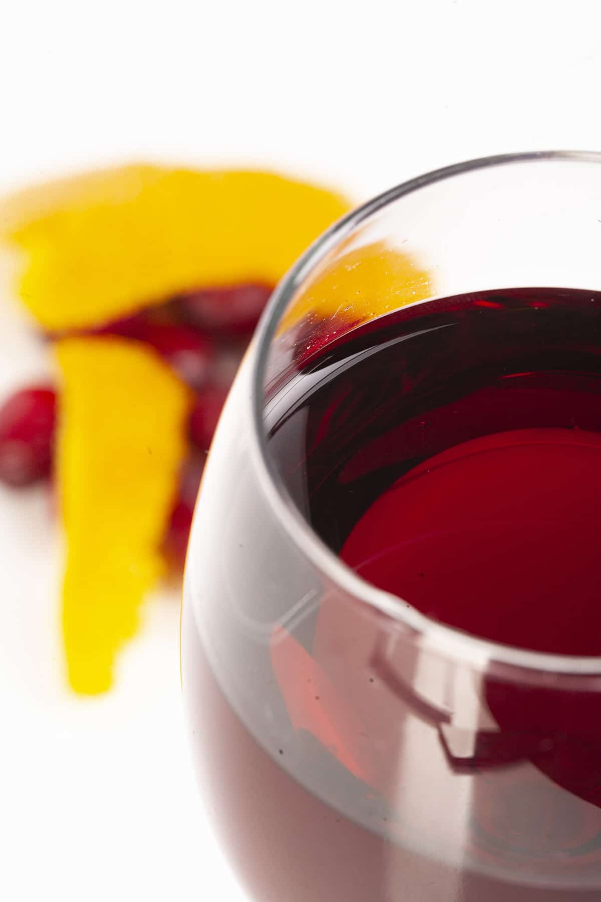 A close up view of a wine glass with with a deep red wine. There are cranberries and orange peels at the base of the glass, against a white background.