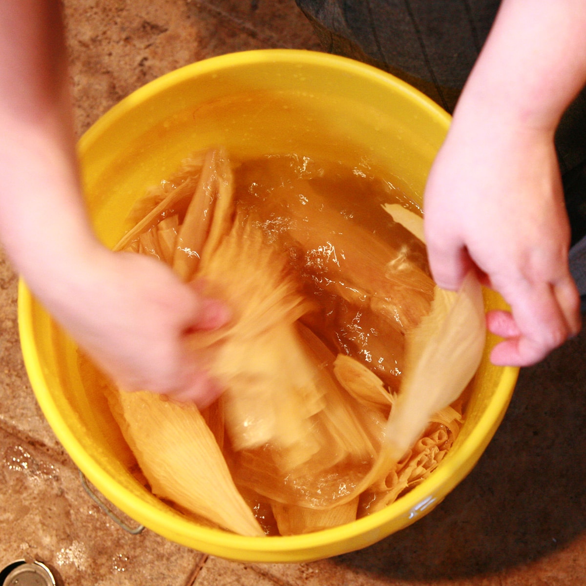 Hands are separating a bag of corn husks, in a large yellow bucket of water.