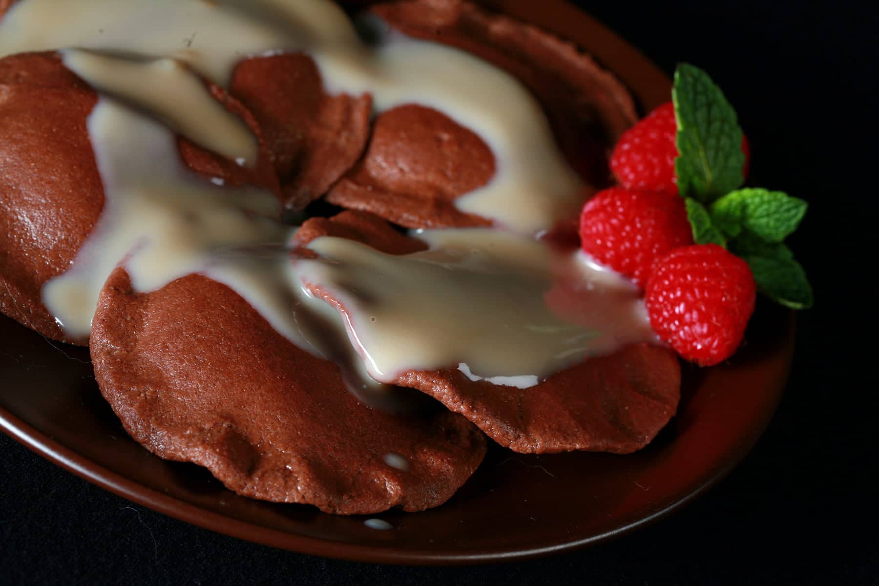 Chocolate Ravioli, arranged on a dark plate. It is drizzled with a cream sauce, and raspberries and a mint spring.