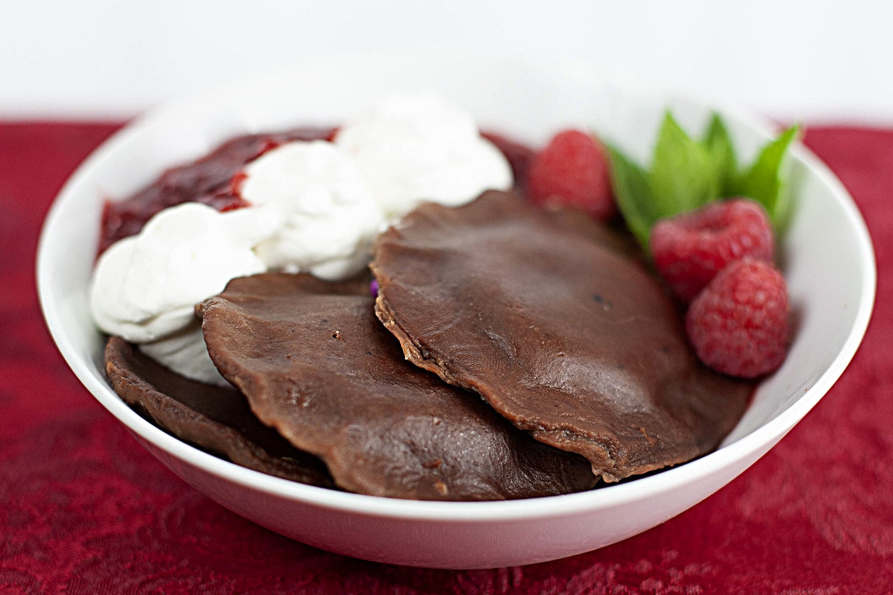 A white bowl holds 2 large chocolate ravioli. There is whipped cream and raspberry sauce on the side, and it is garnished with raspberries and mint.