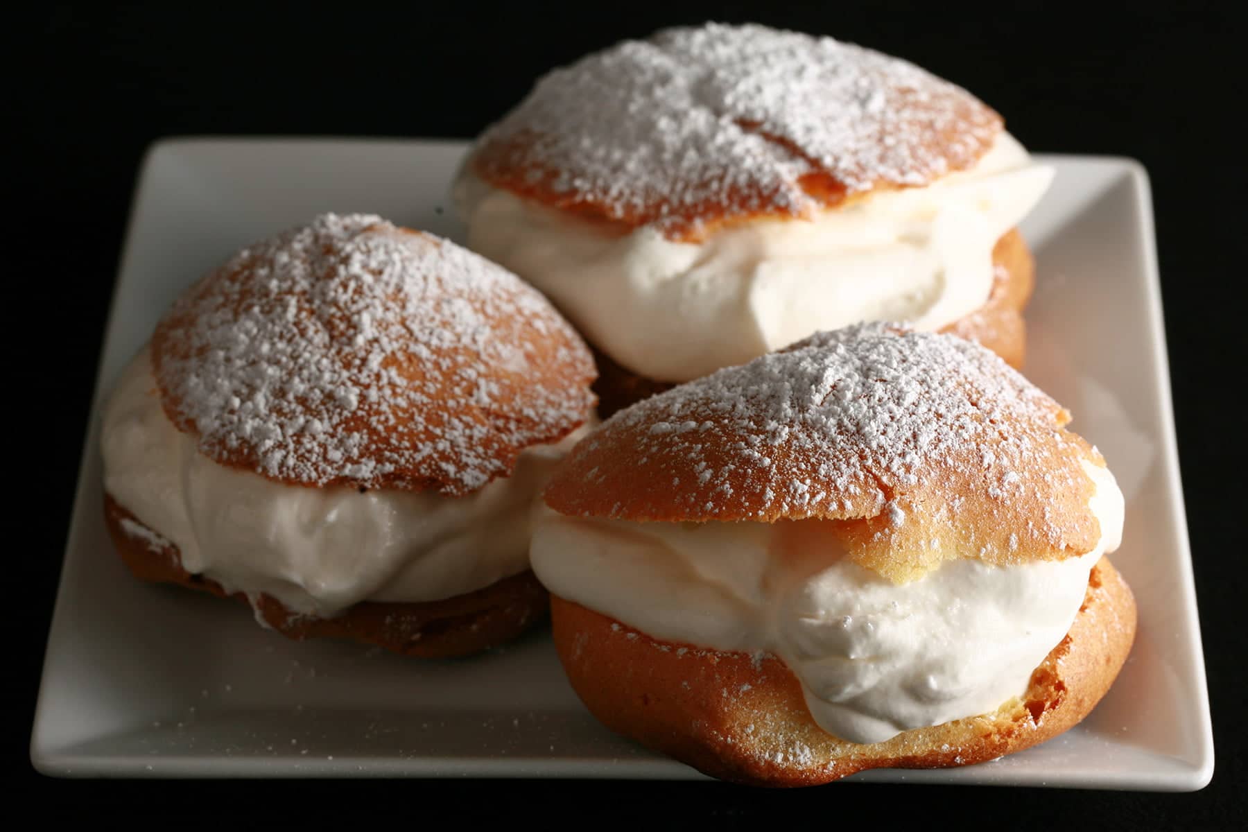 3 Large Cream puffs - with whipped cream in the center, topped with powdered sugar - are pictured on a white plate.