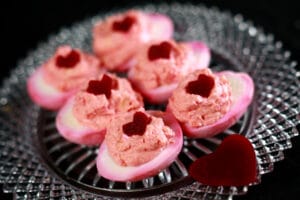 6 pink tie dyed deviled eggs on a glass plate. Each is topped with a heart shaped piece of pickled beet.