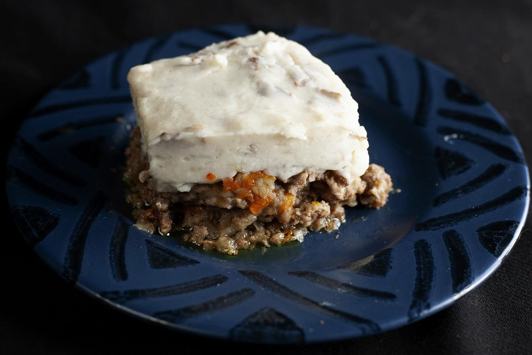 A square serving of shepherd's pie is centered on a blue plate. Carrots and parsnips are visible in the meat/veg base, and it's topped with a thick layer of mashed potatoes.