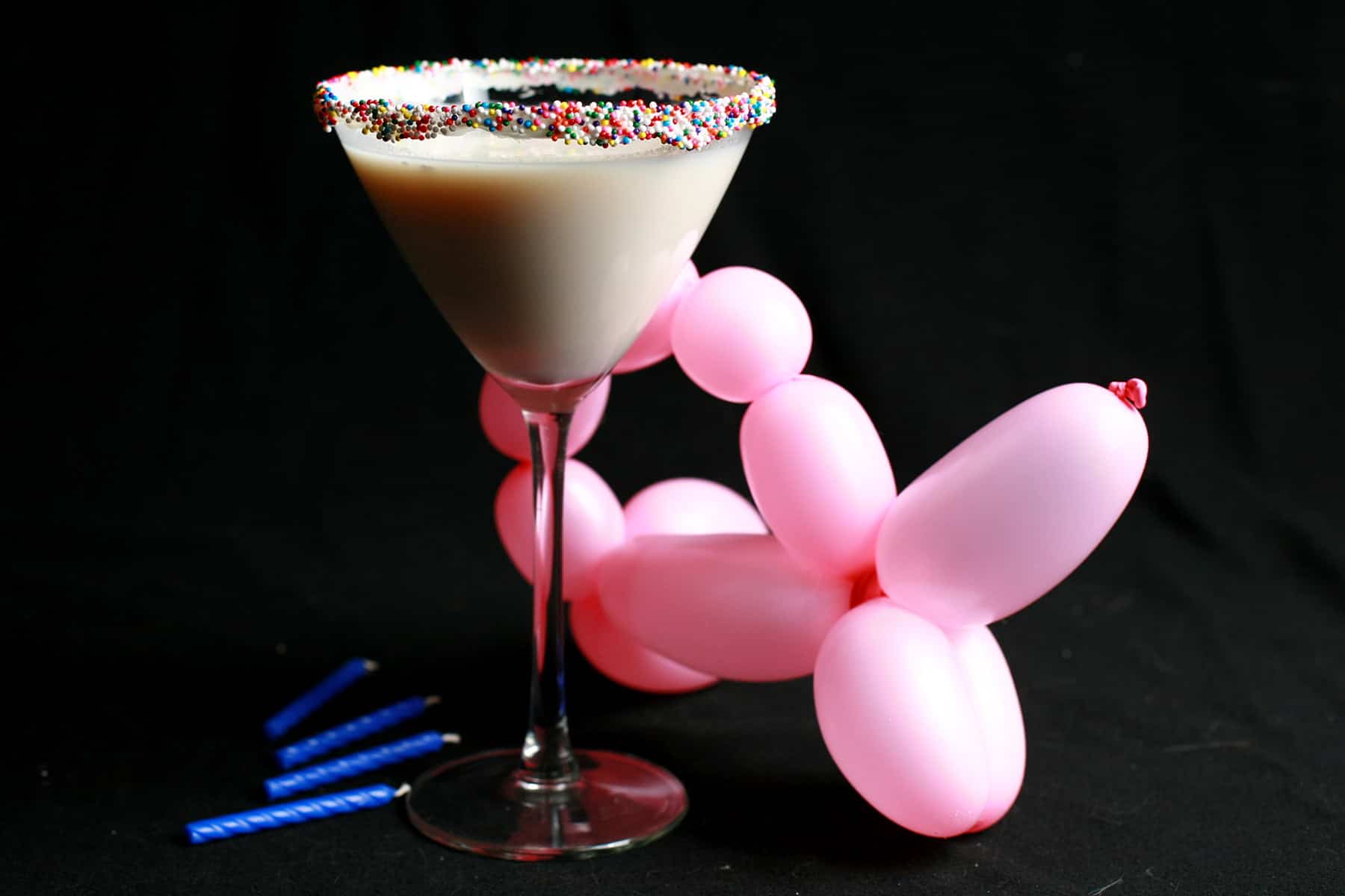 A close up view of a birthday cake martini - a creamy white drink in a martini glass. The rim is coated with colourful nonpareils.  There is a pink ballon animal and blue birthday candles at the base of the glass.