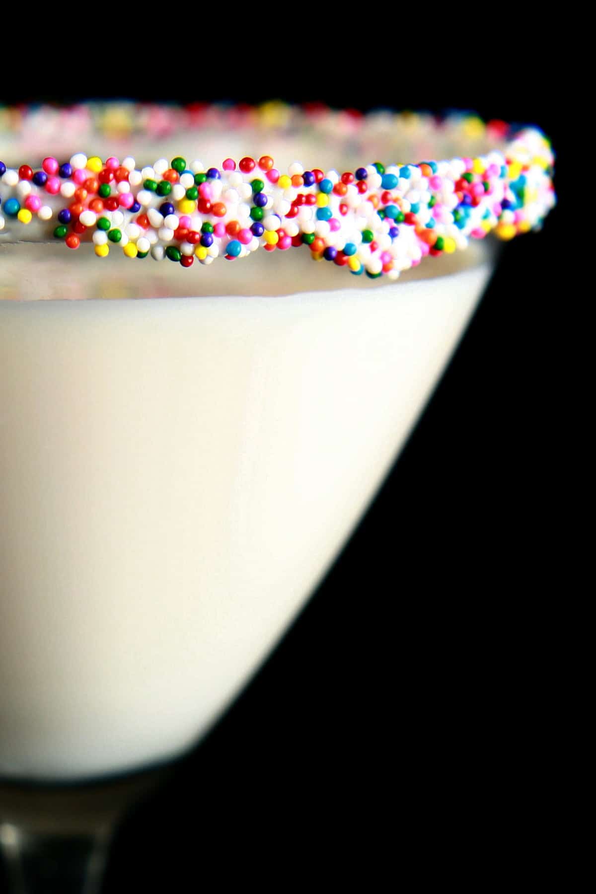 A close up view of a birthday cake martini - a creamy white drink in a martini glass. The rim is coated with colourful nonpareils.