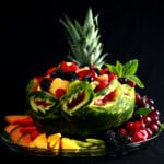 A large, brightly coloured watermelon bowl display. The watermelon is carved with a Caladium Leaf design, and is filled and surrounded by a rainbow assortment of fruit.