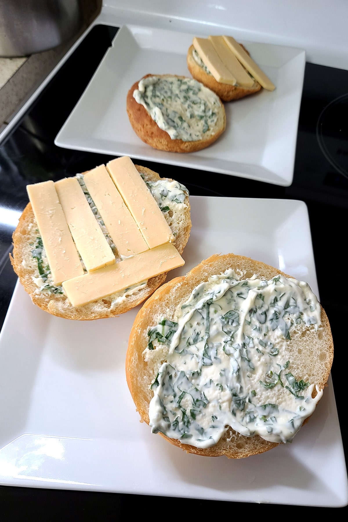 Two square white plates have a bun on each.  Both sides are spread with basil mayo, one side also has cheese slices on it.