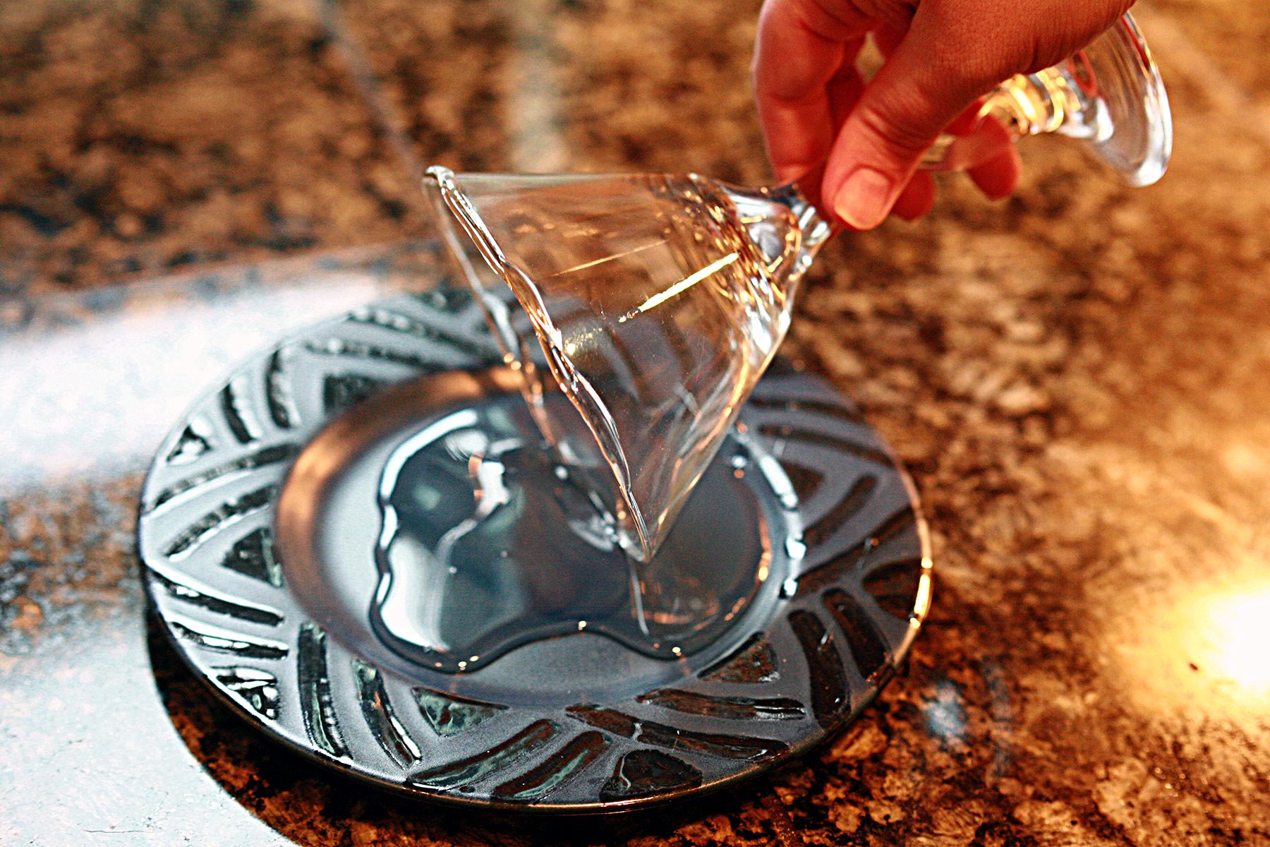 A hand dips the edge of a martini glass in a puddle of clear liquid, on a blue plate.