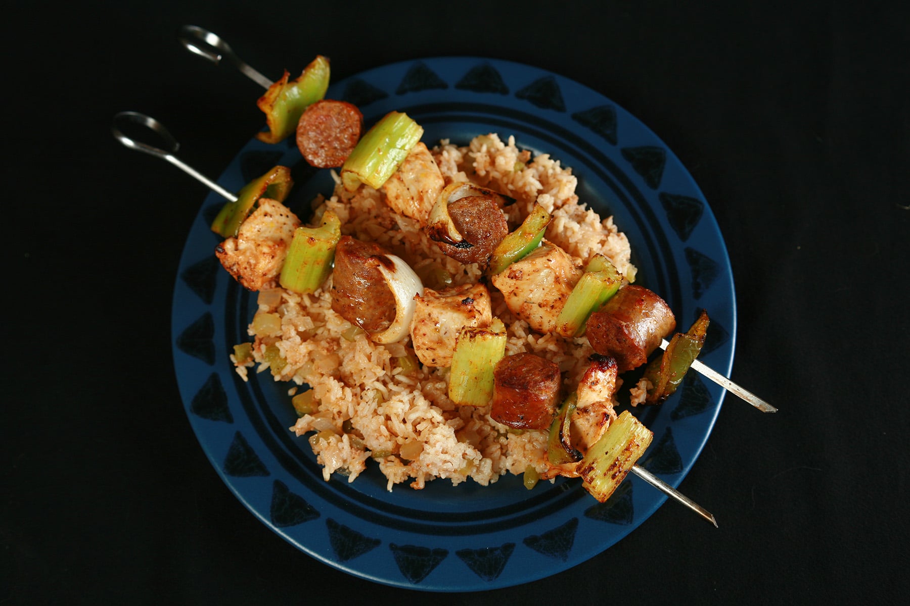 Two grilled skewers of chicken, sausage, green onions, onion, and celery on a bed of intensely seasoned "dirty" rice. rice.