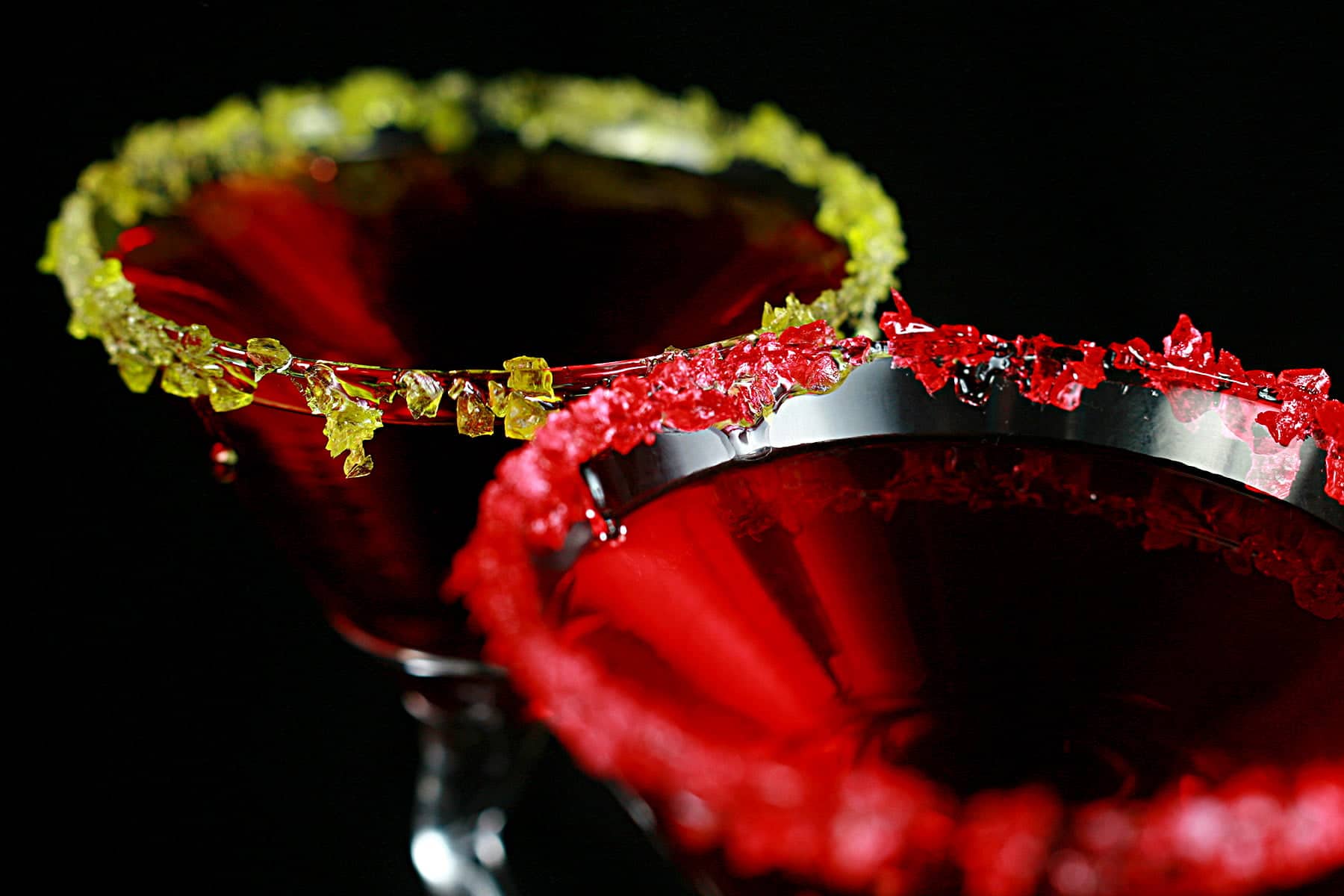 Two Martini cocktails - a Red drink in a martini glass. One is rimmed with crushed green candy, the other is rimmed with a red crushed candy.