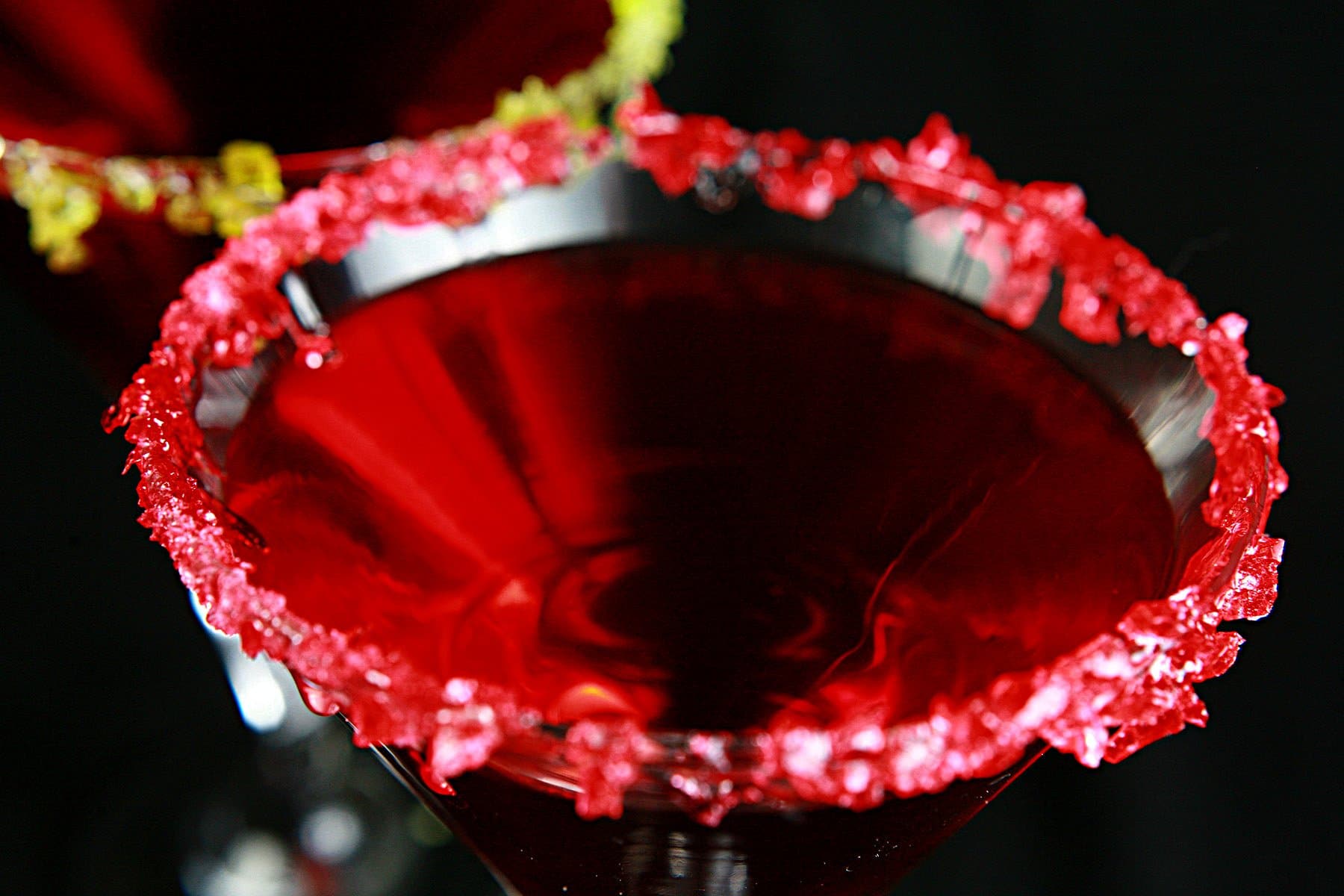 Two Martini cocktails - a Red drink in a martini glass. One is rimmed with crushed green candy, the other is rimmed with a red crushed candy.