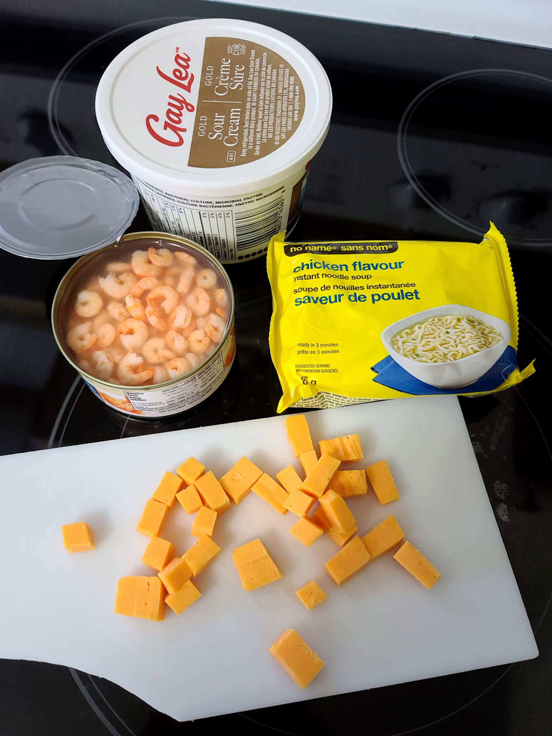 Ramen, a can of shrimp, cheese, and sour cream are laid out on a stove top.