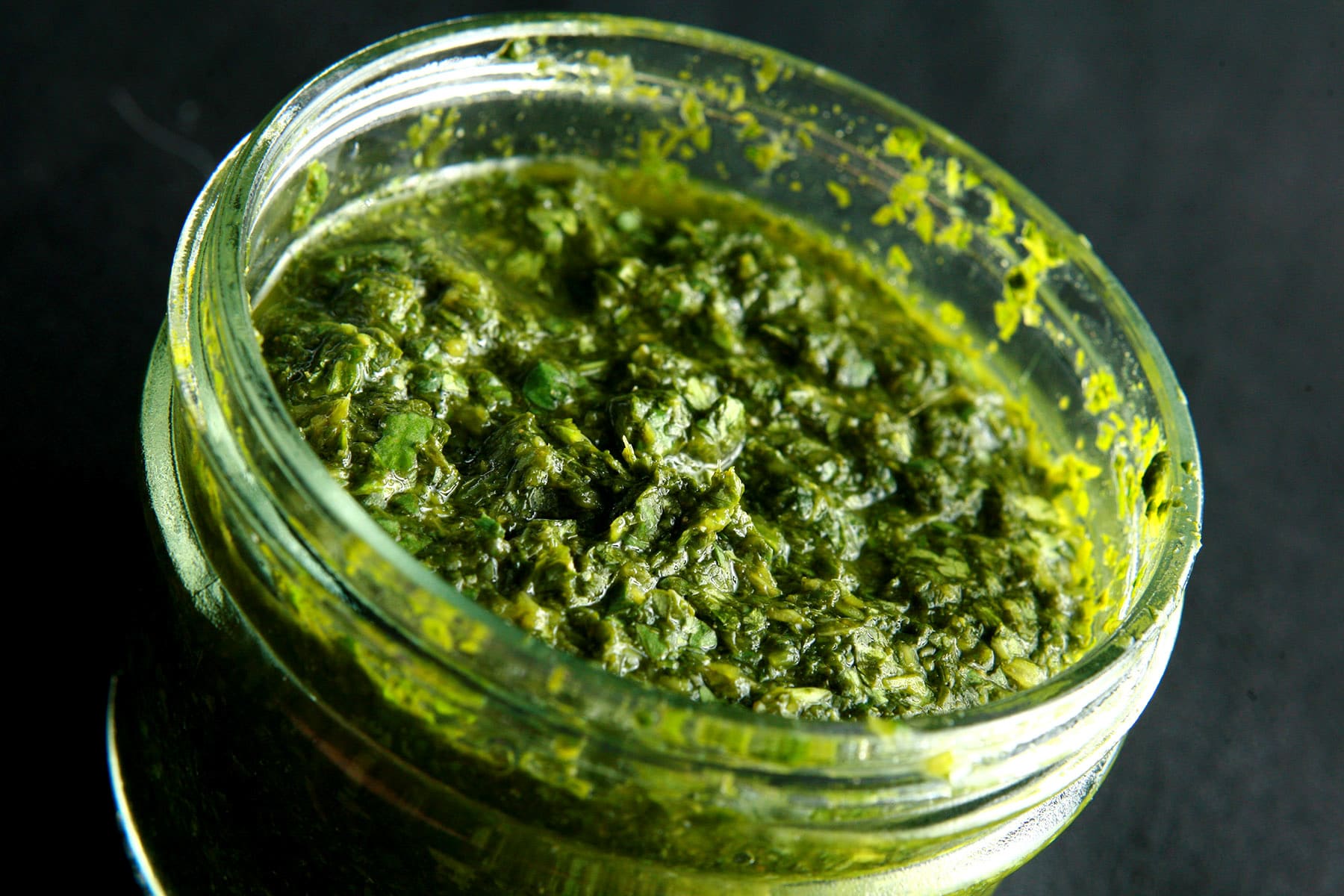 A close up view of a jar of cilantro mint chutney, a deep green dip that looks like pesto.